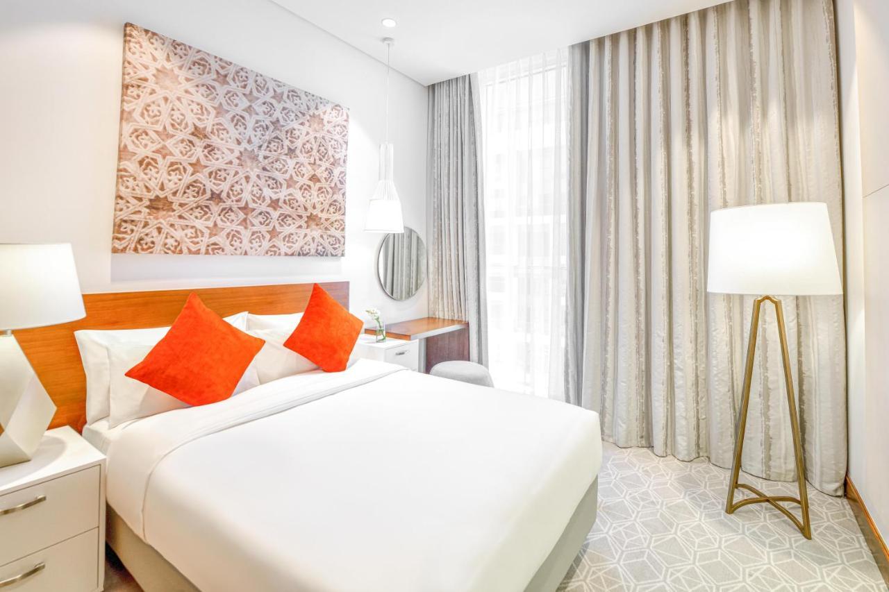 Grand Mercure Dubai City Εξωτερικό φωτογραφία The photo shows a modern hotel room featuring a comfortable queen-sized bed with white linens and orange accent pillows. There is a decorative wall art piece above the bed, adding a stylish touch to the room. A round mirror is mounted on the wall abo