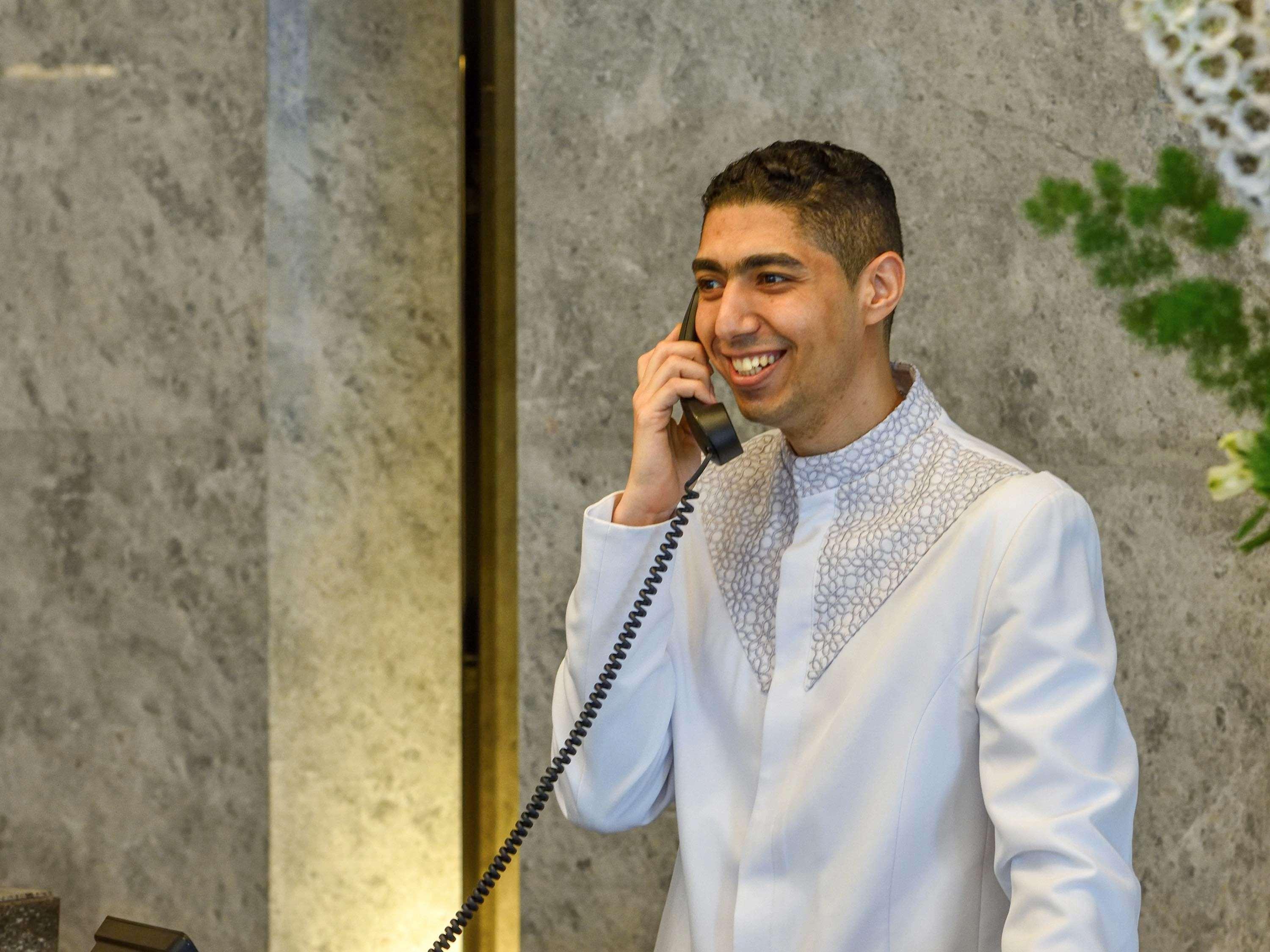 Grand Mercure Dubai City Εξωτερικό φωτογραφία The photo shows a young man dressed in a formal white outfit with decorative details around the collar. He is smiling and is speaking on a vintage-style telephone. The background appears to have a marble texture, and there are some plants or floral a