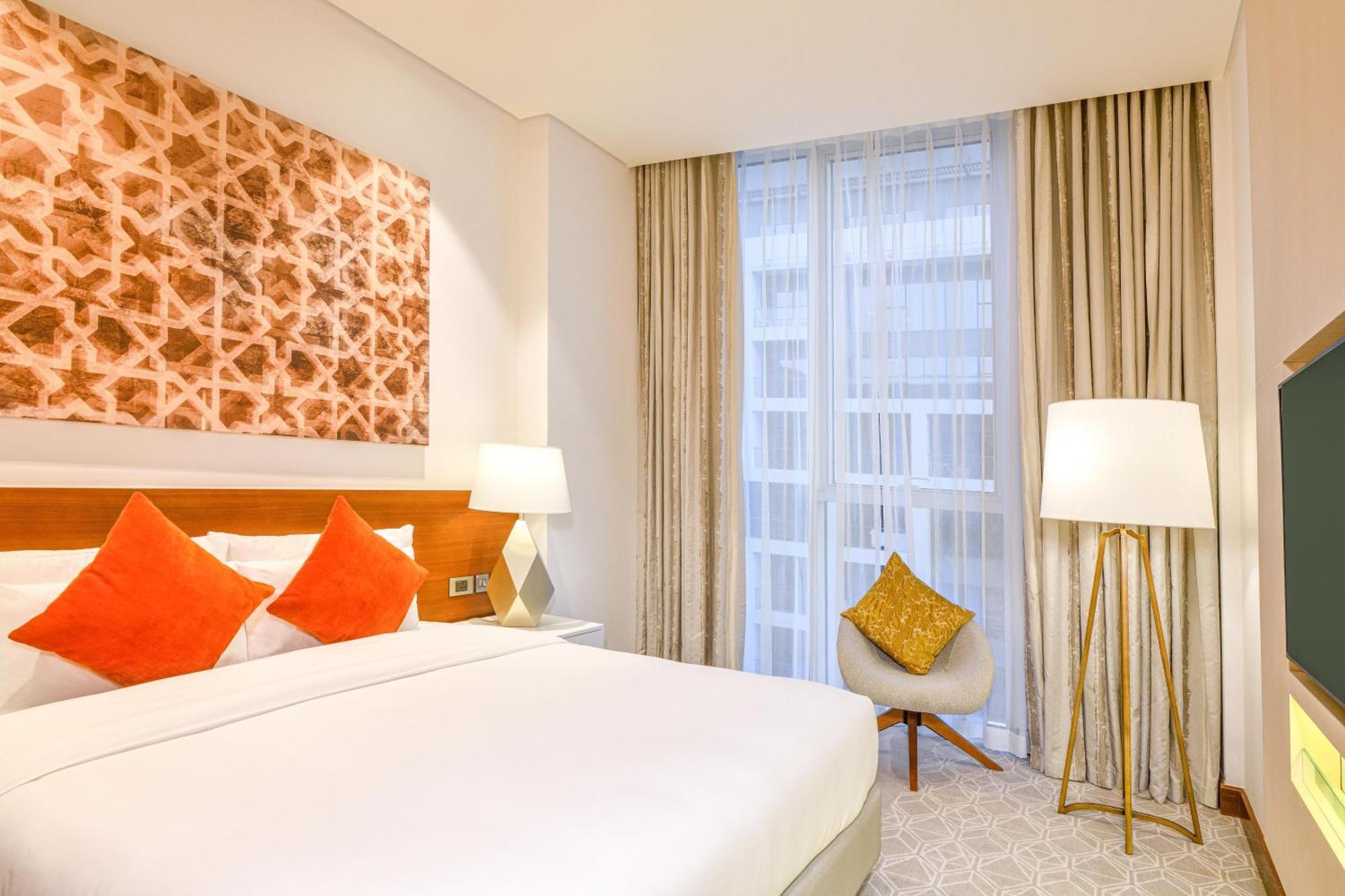 Grand Mercure Dubai City Εξωτερικό φωτογραφία The photo shows a modern hotel room with a cozy and elegant design. There is a large bed with crisp white linens, accompanied by decorative cushions in shades of orange. To the side, there are two bedside lamps providing soft lighting. A colorful pie